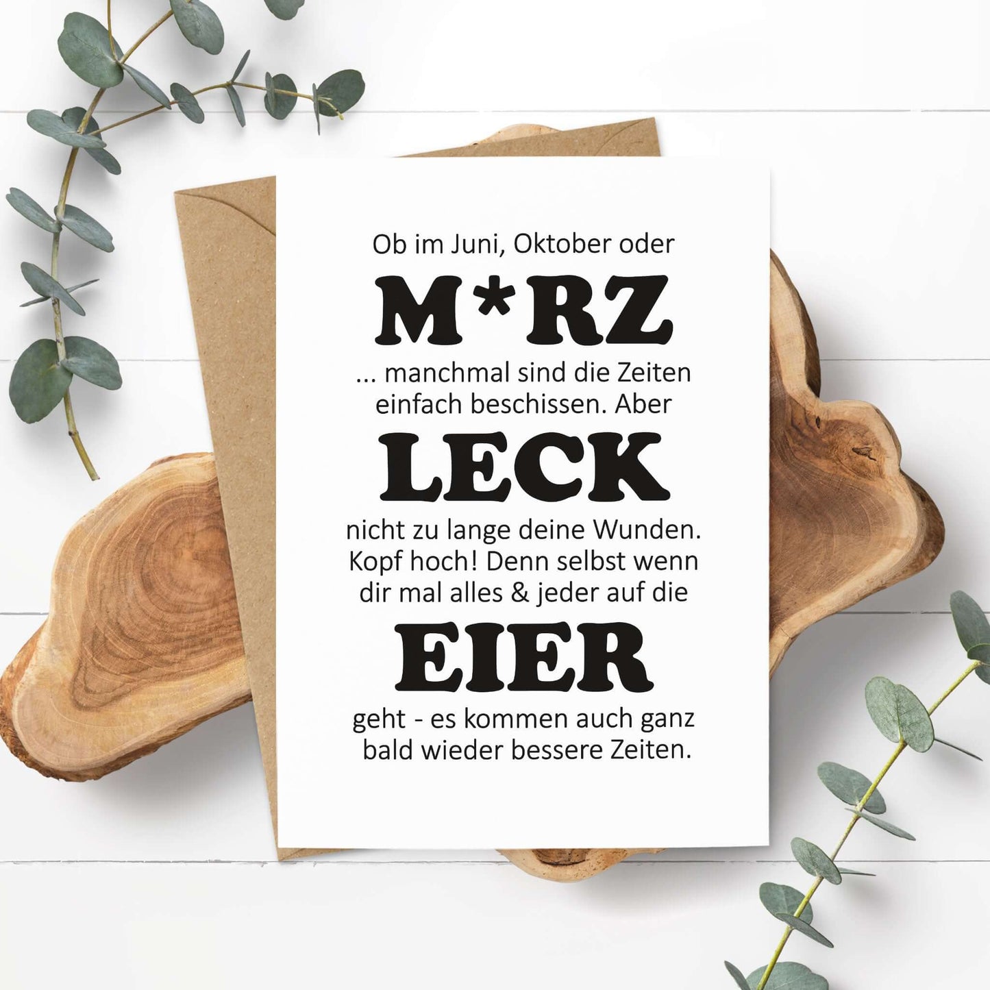 witzige Aufmunterungskarte M*RZ LECK EIER auf Holzuntergrund mit Eukalyptuszweigen.