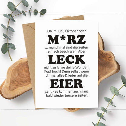 witzige Aufmunterungskarte M*RZ LECK EIER auf Holzuntergrund mit Eukalyptuszweigen.