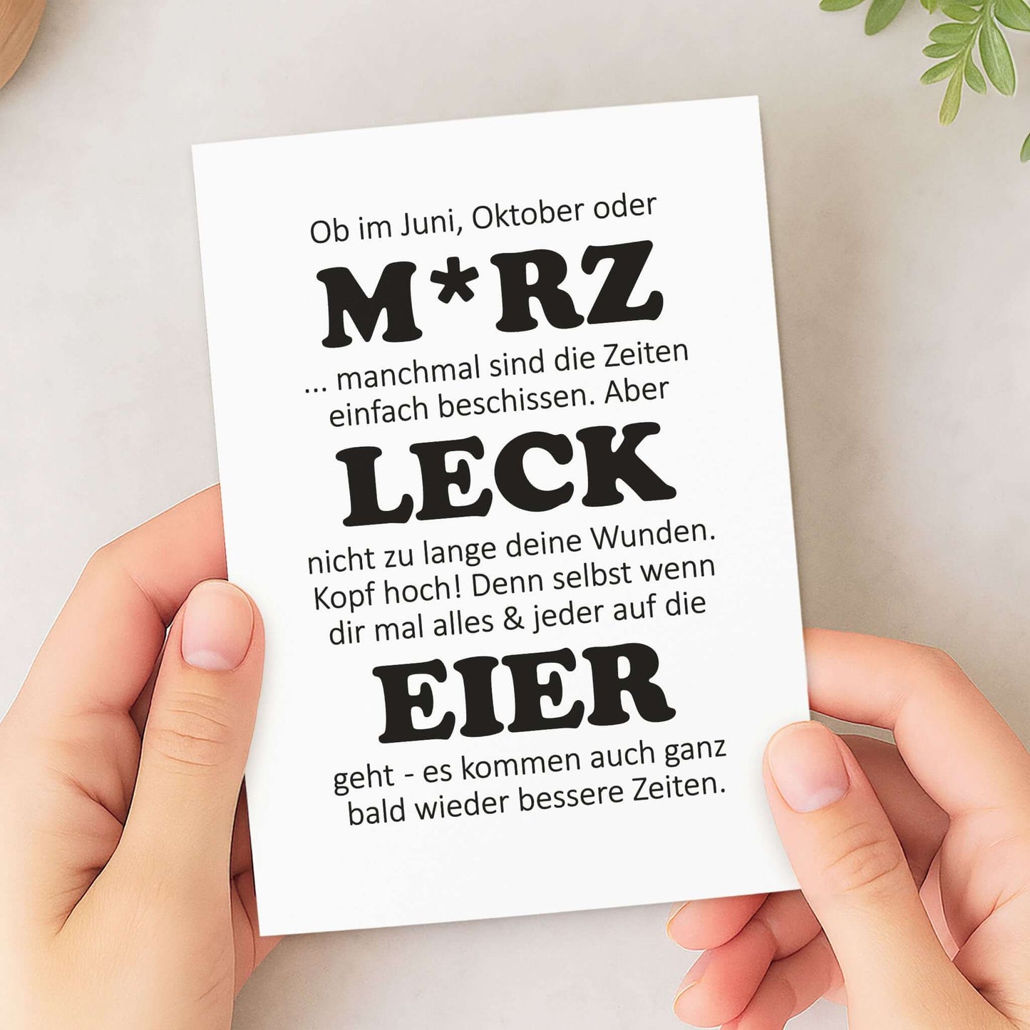 witzige Aufmunterungskarte M*RZ LECK EIER in den Händen einer Person, mit humorvollem Text für Aufmunterung.