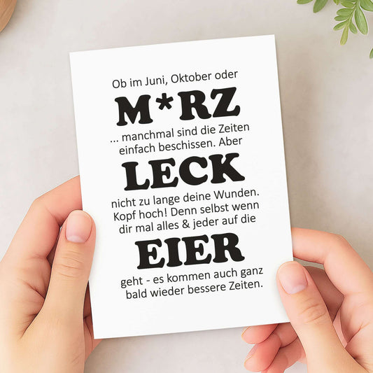 witzige Aufmunterungskarte M*RZ LECK EIER in den Händen einer Person, mit humorvollem Text für Aufmunterung.