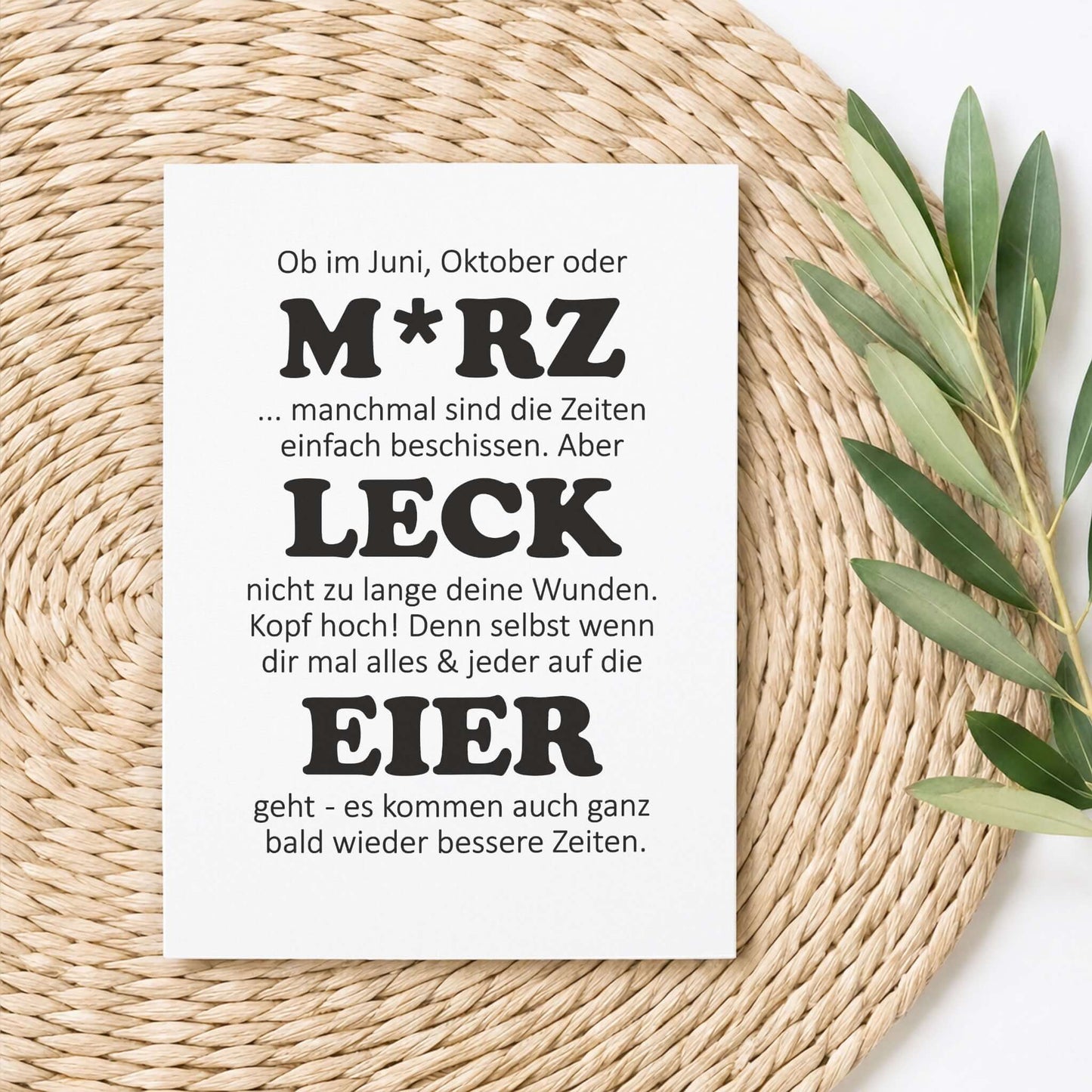 witzige Aufmunterungskarte M*RZ LECK EIER auf einem Tisch mit Pflanzen, humorvoller Spruch auf Postkarte.