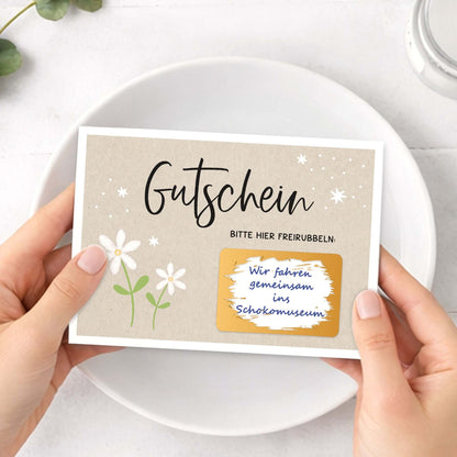 Rubbelgutschein weiße Blumen in Händen mit Gutscheintext und Freirubbelbereich.